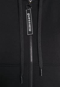 Sweat à capuche noir avec une fermeture éclair visible et des cordons, avec une étiquette noire rectangulaire portant la mention "guess||1981" attachée au curseur de la fermeture.