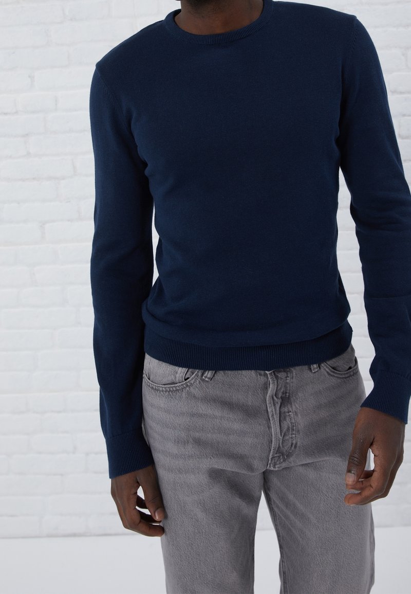 Pull en maille bleu marine avec col rond et poignets côtelés, associé à un jean gris clair. Le pull présente une texture lisse et une coupe confortable.