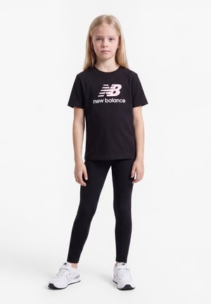 Joven chica con camiseta negra de New Balance, leggings negros y zapatillas blancas de New Balance, de pie sobre un fondo blanco.
