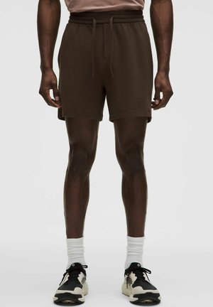 Shorts marron avec une taille élastique et un cordon de serrage, présentant une coupe décontractée. Portés avec des baskets noires et blanches et des chaussettes blanches montantes.