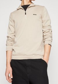 Pull beige avec col à fermeture éclair quart, accents côtelés aux épaules et petit logo noir. Texture douce, silhouette ajustée et manches longues.
