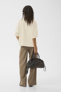 Svart håndveske med krokodillemønster, rund form, dobbelthåndtak og avtagbar stropp, kombinert med en løs beige topp og brune bukser.