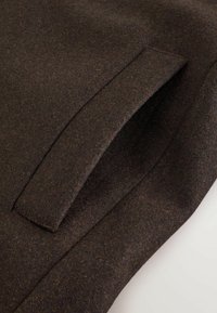 Brauner Wollstoff mit glatter Textur und seitlicher Taschenapplikation. Das Material ist dicht gewebt und weist dezente Farbnuancen auf.