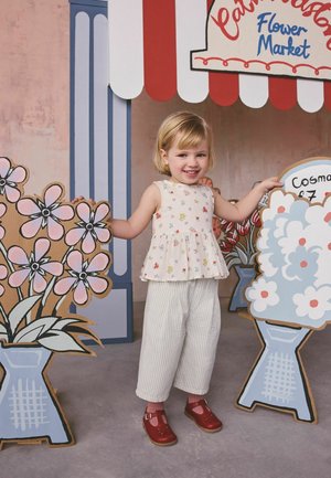 Smilende barn iført blomsterprintet top og stribede bukser står mellem store kartonblomster under en rød- og hvid-blomstret markise.