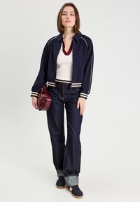 Marineblaues Bomberjacke mit weißen Streifen, cremefarbener gerippter Pullover, dunkle Jeans und burgunderrotes Handtasche; Modell steht vor einem neutralen Hintergrund.