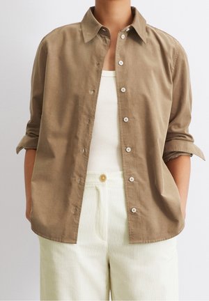 Persoon draagt een lichtbruine blouse met knopen en opgerolde mouwen over een wit hemdje en witte broek met hoge taille.