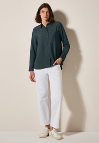 Camisa de botones verde oscuro con mangas largas y un bolsillo en el pecho, combinada con pantalones rectos blancos y zapatillas crema.