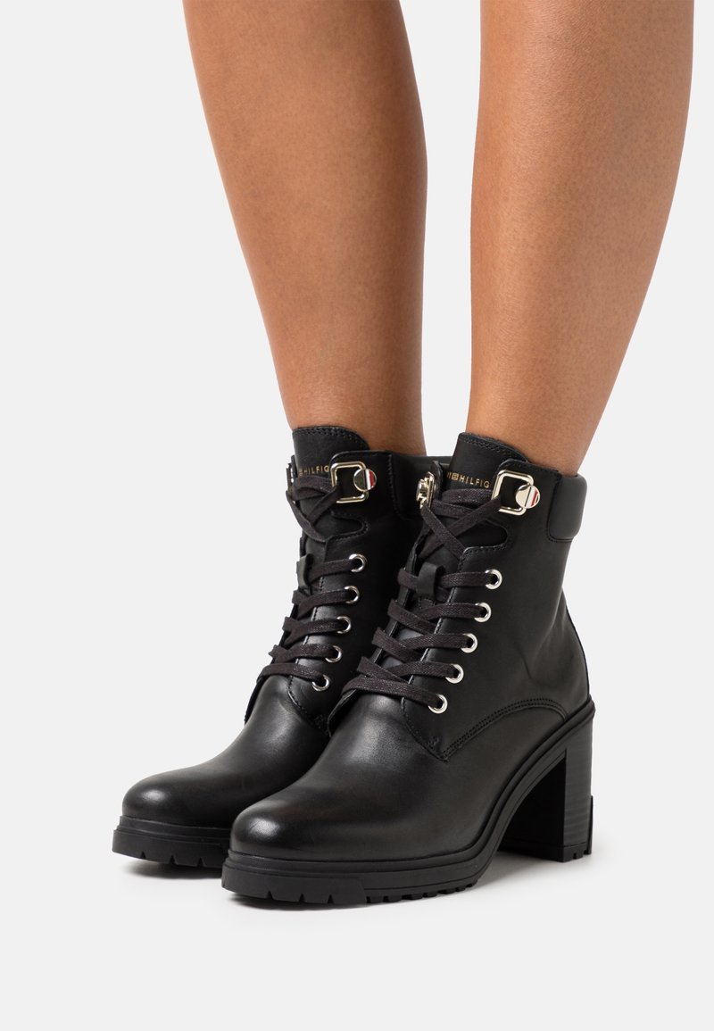 Bottes noires en cuir à lacets, à talons blocs, portées sur des jambes nues sur un fond neutre.