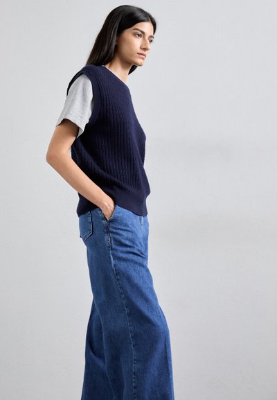 Marineblå ribbestrikket ermela genser over en lysegrå T-skjorte, kombinert med vide blå denimjeans. Glatt tekstur, avslappet passform.