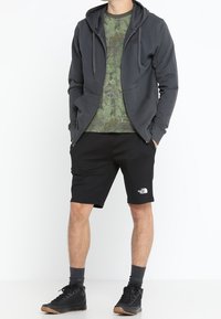 Felpa grigia con zip sopra una maglietta verde a mimetica, abbinata a pantaloni corti neri. Scarpe nere e calze grigie completano l'outfit.