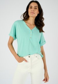 Camisa de botones de color turquesa claro con escote en V y mangas cortas dobladas, combinada con jeans blancos. Textura de tela suave y lisa.