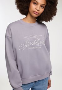 Jasnoszara bluza z okrągłym dekoltem i długimi rękawami. Posiada biały napis "Stockholm Leisure Club 1985". Miękka faktura.