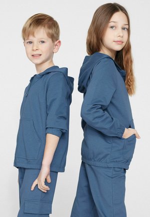 Zwei Kinder stehen Rücken an Rücken und tragen passende blaue Kapuzenpullover und Hosen vor einem schlichten weißen Hintergrund.