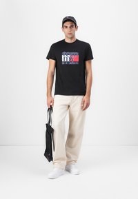 T-shirt en coton noir avec un logo graphique, pantalon large beige, baskets blanches et un sac noir, se tenant devant un fond blanc.