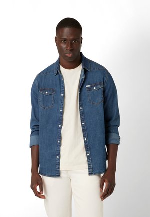 Denim shirt in dark blue with button-front closure, two chest pockets, and traditional collar; paired with a light t-shirt and white pants.