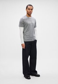 Mężczyzna stoi z rękami w kieszeniach, nosząc szary t-shirt Calvina na białej koszuli z długim rękawem, ciemne szerokie dżinsy oraz czarne buty na jednolitym tle.