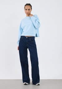 Sweatshirt azul claro, cropped, com mangas largas, combinado com jeans de cintura alta azul escuro. Ténis com detalhes em preto e branco visíveis.