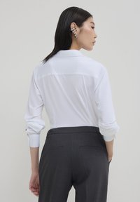 Donna con capelli neri lisci, che indossa una camicia bianca a maniche lunghe piegata, pantaloni grigio scuro e diversi orecchini a cuffia in argento, vista di spalle.