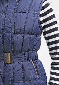 Marinblå puffer väst med horisontell quiltad design, sidoficka med dragkedja och guldfärgad spänne vid midjan, lager på lager över en svart- och vitrandig topp.