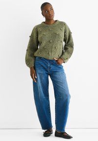 Grüner strukturierter Pullover mit Bobbel-Details, kombiniert mit lockeren blauen Jeans und schwarzen Ballerinas. Das Model posiert mit einer Hand auf der Hüfte vor einem weißen Hintergrund.