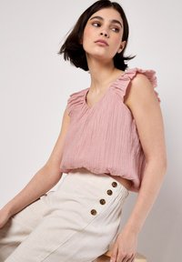 Rosa V-Ausschnitt-Oberteil mit rüschierten Schultern, strukturiertem Stoff, kombiniert mit hochtaillierten weißen Hosen, die vier braune Knopfakzente aufweisen.