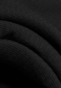 Close-up van zwarte stof met een gestructureerd breipatroon, gevouwen in zachte golven, waarbij het gedetailleerde stikwerk en de diepte van het materiaal worden benadrukt.