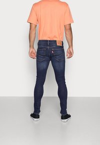 Jeans de mezclilla azul oscuro con un diseño ajustado, presentando un parche de cuero con el logo en la cinturilla y bolsillos traseros estándar. Combinados con una camiseta naranja.