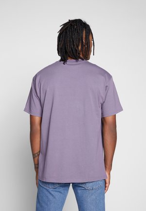 Person mit dunklen Dreadlocks, die ein schlichtes lila Kurzarm-T-Shirt und blaue Jeans trägt, von hinten vor einem grauen Hintergrund dargestellt.