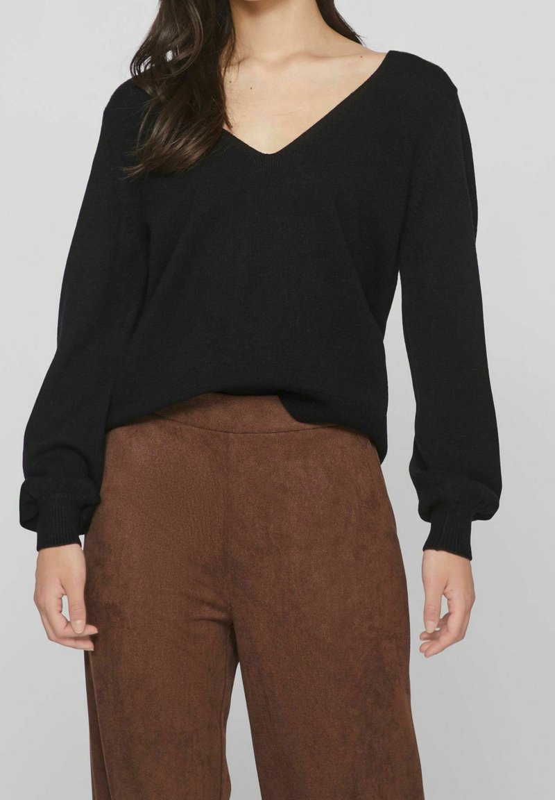 Femme portant un pull noir à col en V et manches longues ainsi qu'un pantalon marron taille haute, debout avec les bras détendus le long du corps.