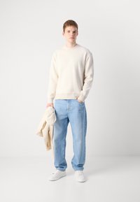 Helles beigefarbenes, geripptes Sweatshirt, blaue Jeans, hält eine hellbeige Jacke, weiße Sneakers, steht vor einem schlichten weißen Hintergrund.