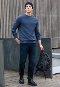 Jersey azul texturizado, jeans oscuros, botines negros y una mochila negra sostenida en la mano. Fondo de madera natural con detalles en gris.