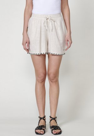 Linned shorts i lys beige med elastisk talje, snøre og sort dekorativ kant langs kanten. Båret med sorte remsandaler.