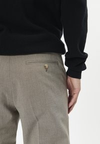 Beige, sima textúrájú, testreszabott nadrág, hátul gombos zsebbel, fekete texturált pulóverrel viselve.