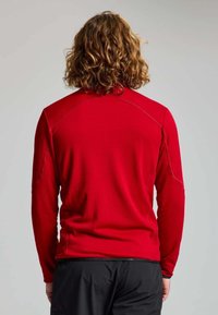 Personne avec des cheveux bruns bouclés portant une chemise rouge à manches longues et un pantalon noir, vue de dos sur un fond uni.