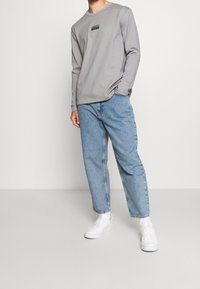 Maglietta grigia a maniche lunghe con una piccola toppa nera, abbinata a jeans in denim azzurri a vestibilità rilassata e sneakers bianche.