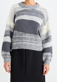 Pull en maille à encolure ronde, présentant des rayures horizontales dans des tons de gris et de crème. Manches volumineuses et détail à l'ourlet raccourci.