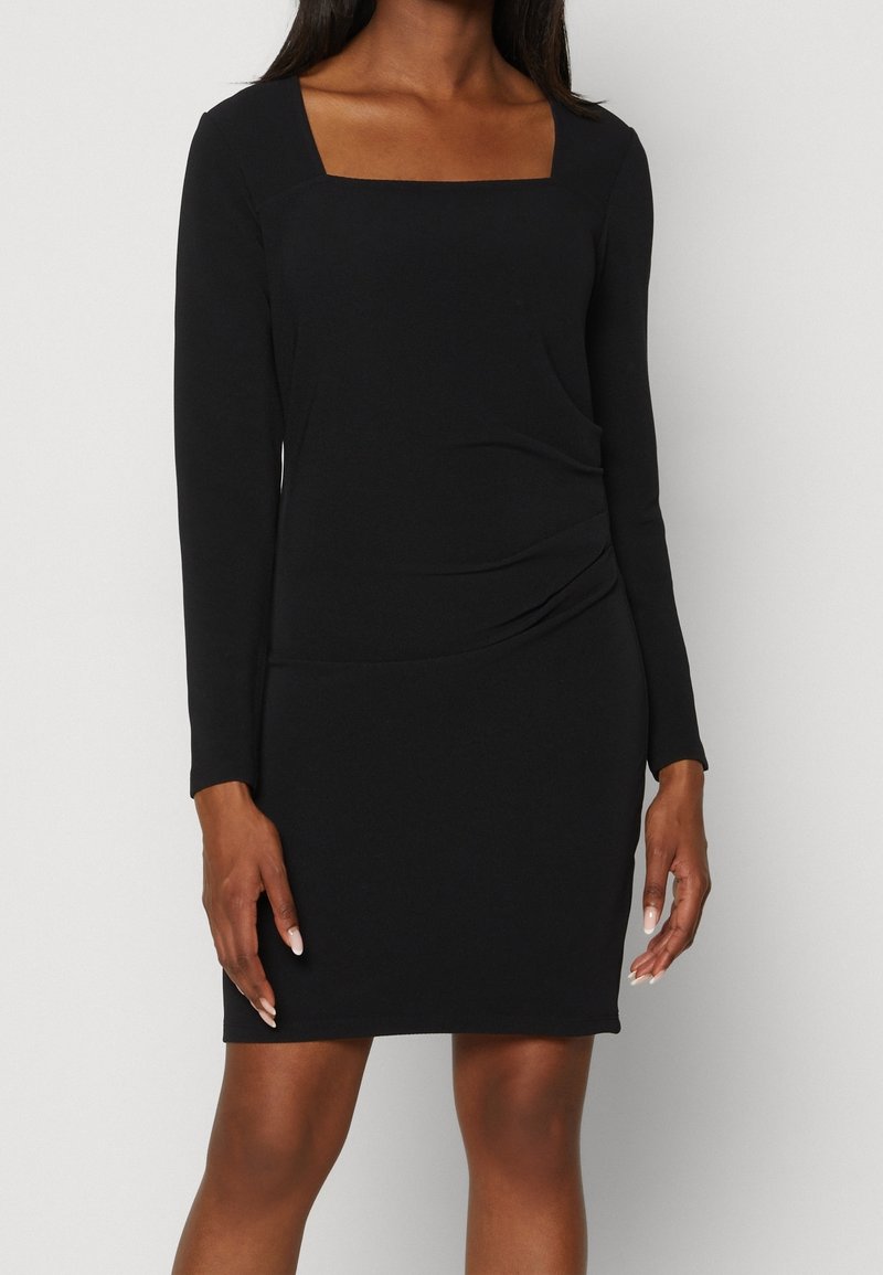 Femme portant une robe noire ajustée à manches longues, avec un décolleté carré et un léger froncé sur le côté, debout devant un fond blanc.