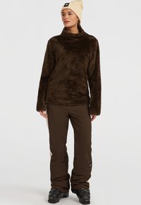 Brun, fluffy fleece pullover med høj krave; parret med brune, afslappede bukser og sorte sneboots. Personen har en cremefarvet hue på.