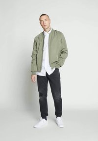 Uomo in piedi con una mano in tasca, indossando una giacca bomber verde, una camicia bianca con bottone, pantaloni neri e sneakers bianche su uno sfondo neutro.