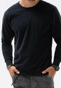 Maglione nero a maniche lunghe con collo rotondo realizzato in cotone, caratterizzato da una texture liscia e polsini e orlo a coste, abbinato a pantaloni grigi.