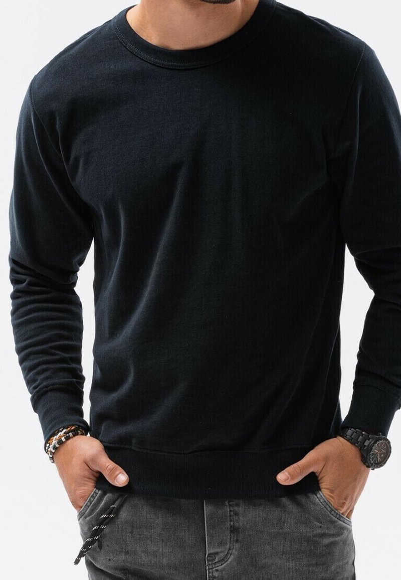 Maglione nero a maniche lunghe con collo rotondo realizzato in cotone, caratterizzato da una texture liscia e polsini e orlo a coste, abbinato a pantaloni grigi.