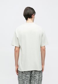 Bărbat văzut din spate, purtând un tricou simplu de culoare gri deschis, supradimensionat, și pantaloni lejeri cu model alb-negru, pe un fundal neutru.