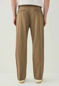 Brown trousers with a straight-cut design, featuring two back pockets with button closures. Smooth texture and tailored fit, paired with white soles.