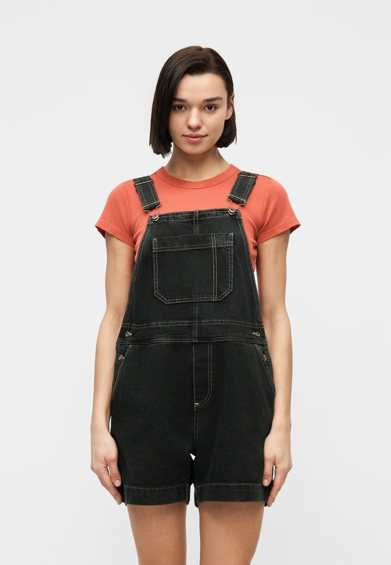 Νεαρή γυναίκα που στέκεται με το πρόσωπο προς τα εμπρός, φοράει κοραλλί κοντομάνικο πουκάμισο και μαύρο κοντό τζιν φορμάκι, μπροστά σε ένα απλό ανοιχτό φόντο.