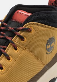 Bota de senderismo impermeable marrón con un upper texturizado, acentos en negro, cordones contrastantes y un logo de árbol en el costado. Etiqueta roja en el interior que muestra la marca.