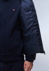 Marineblå lynlåstrøje med bomuldsyderside og quiltet satinfor. Har ribbede manchetter og kant samt et lille logo på brystet.