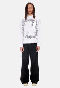 Άτομο με μακριές πλεξούδες ντραντλoκ, φοράει άσπρο πουκάμισο με μαύρο αφηρημένο μοτίβο και φαρδιά μαύρη παντελόνα, στέκεται μπροστά σε απλό φόντο.