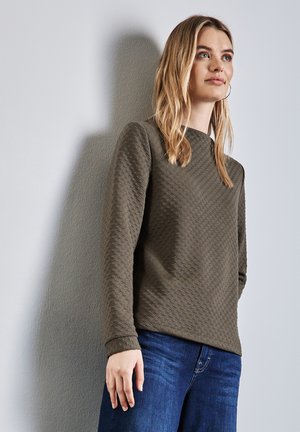 Sweatshirt texturé vert olive avec un col rond, présentant un motif en losanges, assorti à un jean bleu. Tissu lisse, design légèrement ajusté.