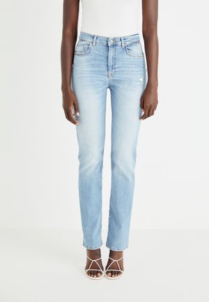 Persoon die lichtblauwe high-waist skinny jeans met lichte slijtage draagt en witte gestripte hoge hak sandaaltjes, staand tegen een witte achtergrond.