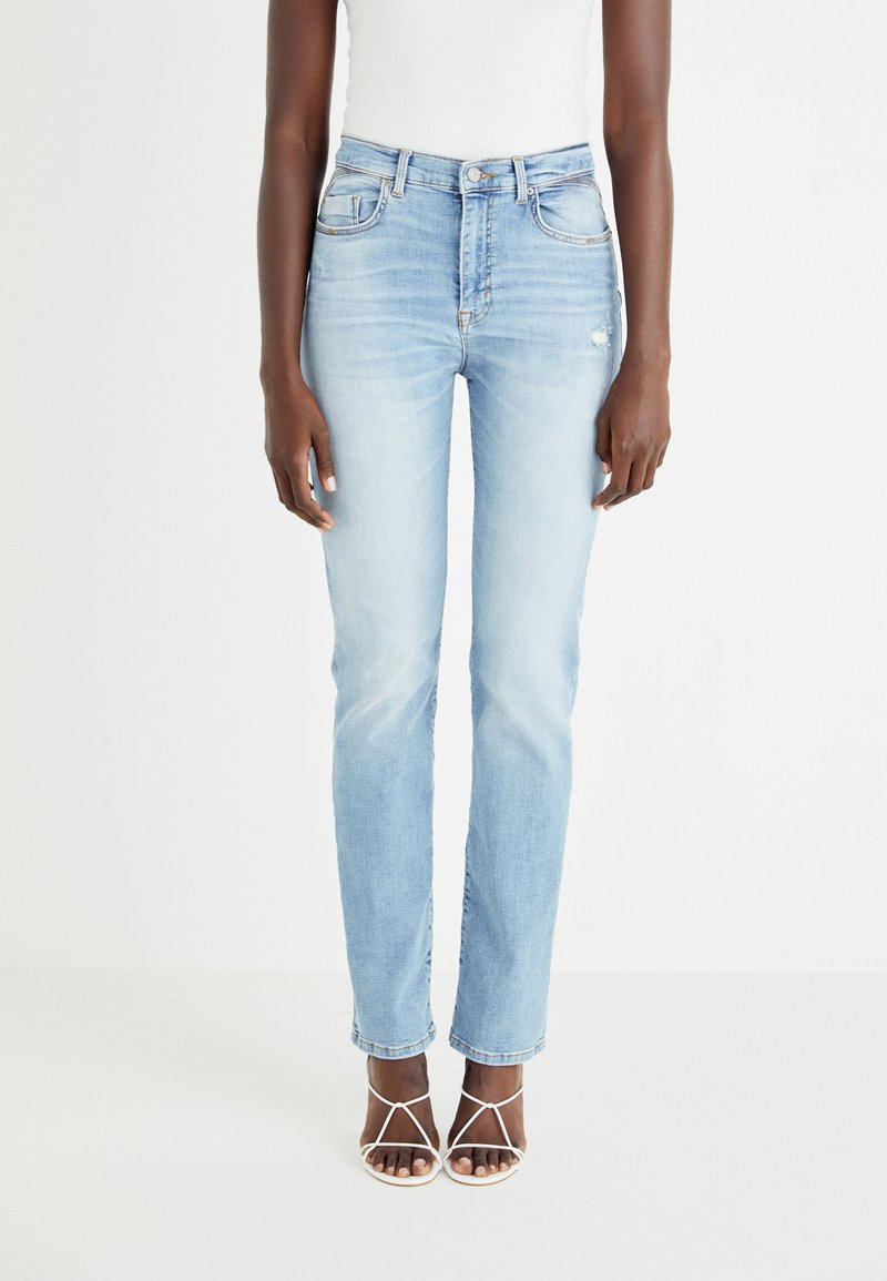 Personne portant un jean skinny bleu clair taille haute avec légère usure et des sandales blanches à talons hauts à brides, debout devant un fond blanc.